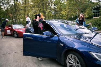 四名十大网络娱乐平台注册的学生爬进了两辆特斯拉汽车的驾驶座和乘客座, 前景是一辆蓝色轿车，背景是一辆运动型红色跑车.