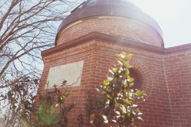 博斯韦尔天文台图片, 一座红砖建筑，顶部有一个铜圆顶，可以打开和关闭，以便观看天空. 大楼上的一块牌匾上写着“博斯韦尔天文台1883”.“这张照片是从一个低角度拍摄的，前景中有一些植物，上面隐约可见建筑. 